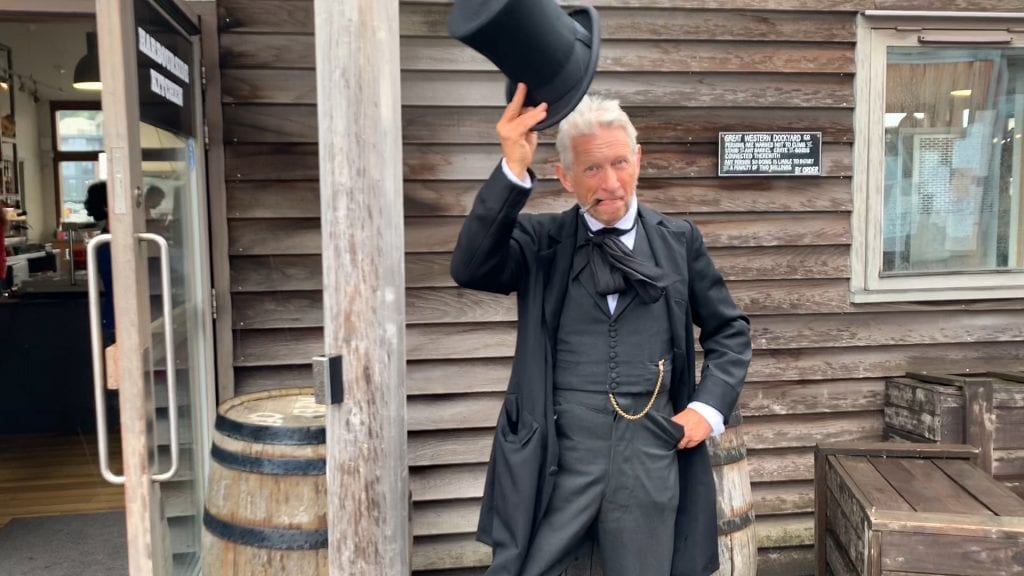 Man dressed as Isambard Kingdom Brunel in black frock coat, waistcoat with fob watch, trousers and black cravat, raising his top hat with a cigar clamped in his teeth. He is standing in front of a wooden building.