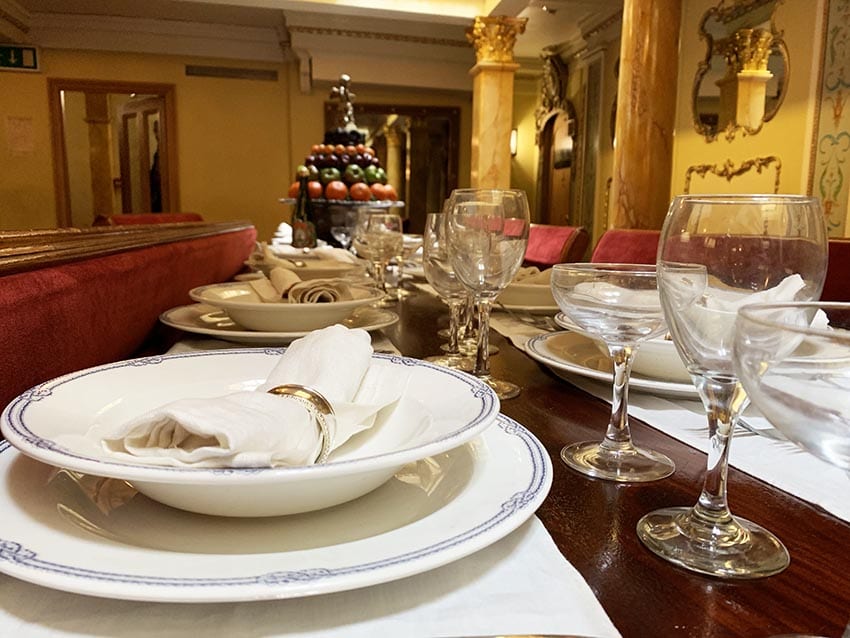 Table set with blue rimmed plates with white napkin in a gold napkin ring and wine and champagne glassed with a display of fruits piled in the background.