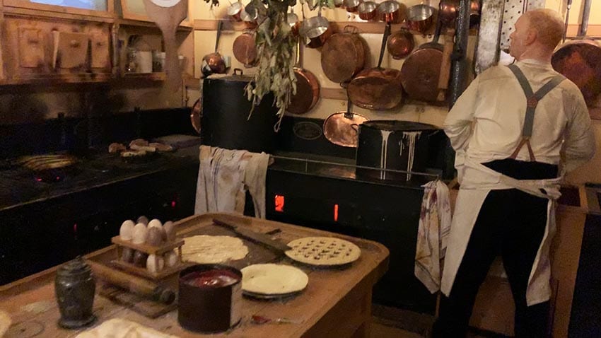 Old fashioned kitchen on the SS Great Britain, with copper pans hanging on the walls. Eggs on the counter and pies being made. The cook has an apron around his waist and is wearing braces.