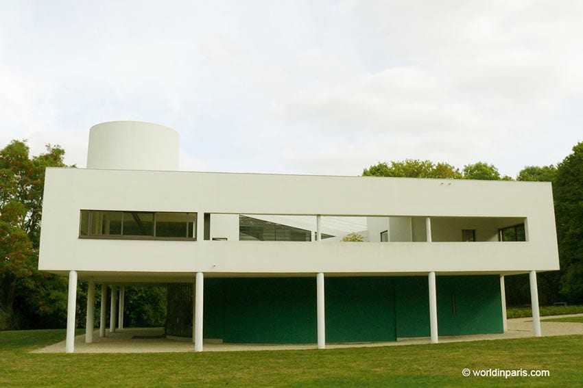 Villa Savoye a modern looking building with white rectangular box with rectangular windows supported on 5 thing columns with a round white tower in the left hand background. 