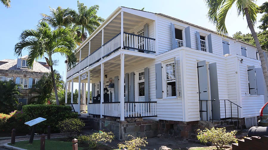White clapperboard two storey house with powder blue shutters. There is a verandah on each floor. Palm trees are in the gardens