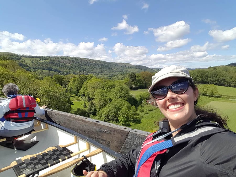 Guide to 17 Amazing UNESCO Heritage Sites in the UK 6 Girl wearing white hat and sunglasses smiling at the camera as she travels in a white boat along the Pontcysyllte Aqueduct with green trees and fields below.