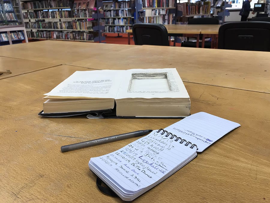 Geocaching, how to be part of the worldwide treasure hunt! 3 open book with a portion cut out with a note pad and pen beside it. In the background are library shelves full of books and chairs and tables