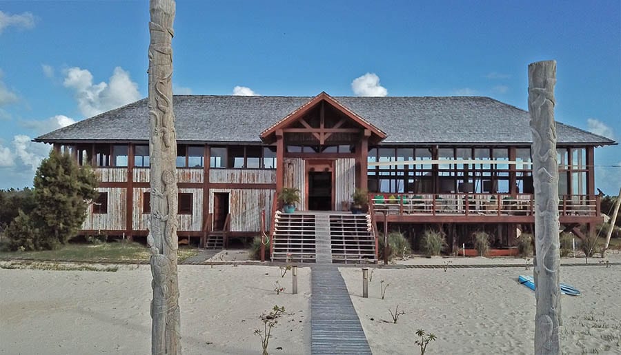 Barbuda – What you Need to Know Before You Go 8 Wooden building with steps going up to it and long, tall glass windows. Two wooden poles with carvings of lizards are either side of the wooden walkway. Barbuda Belle Resort