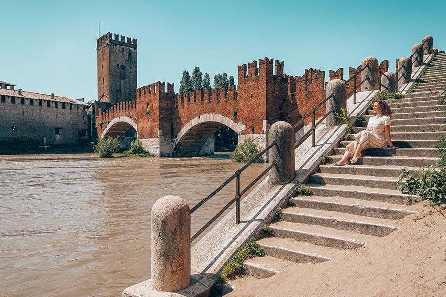 25 of the Best Castles in Italy 10 smiling woman sitting on steps with Verona Castle in red brick with arches to her left