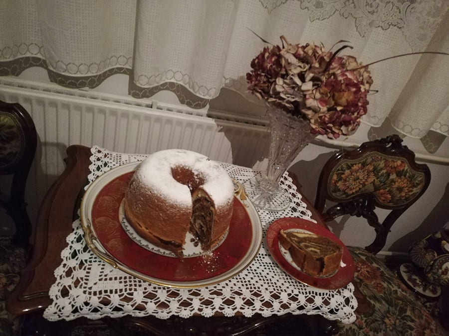 round marbled potica cake from Slovenia dusted with icing sugar and sitting on a red plate. The table has a lace tablecloth and there is dried flowers and net curtains in the background