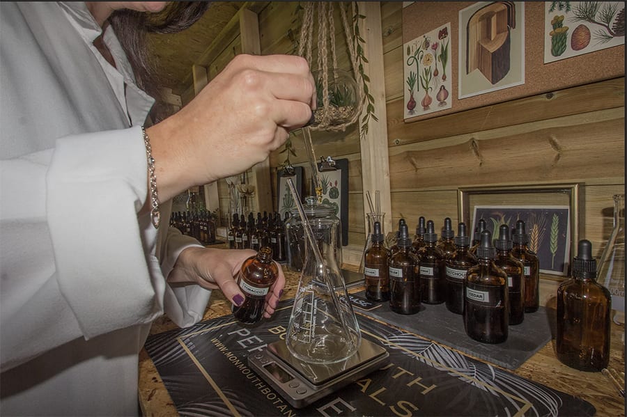 Woman in lab coat measuring out fragrances into a jar on scale from a brown bottle with a row of brown bottles in front of her and botanical prints on the wall
