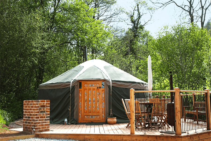 7 of the Best Staycations in Wales & Northern Ireland 6 Yurt with traditional door sitting on a wooden patio with table, chairs and umbrella with trees in the background