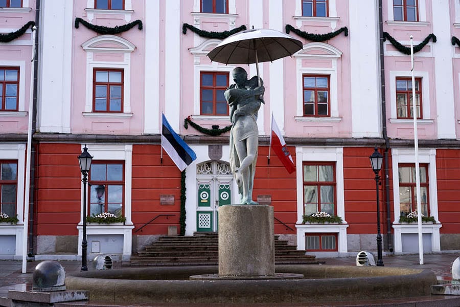 Tartu Town Hall Square with pink and red building behind a fountain with a sculpture of a couple wrapped around each other kissing with the man holding an umbrella above them