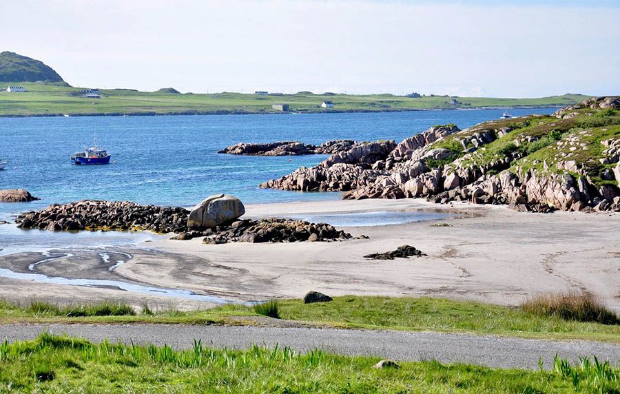 beach with cliffs to one side and boat on the sea. On the far shore are white houses