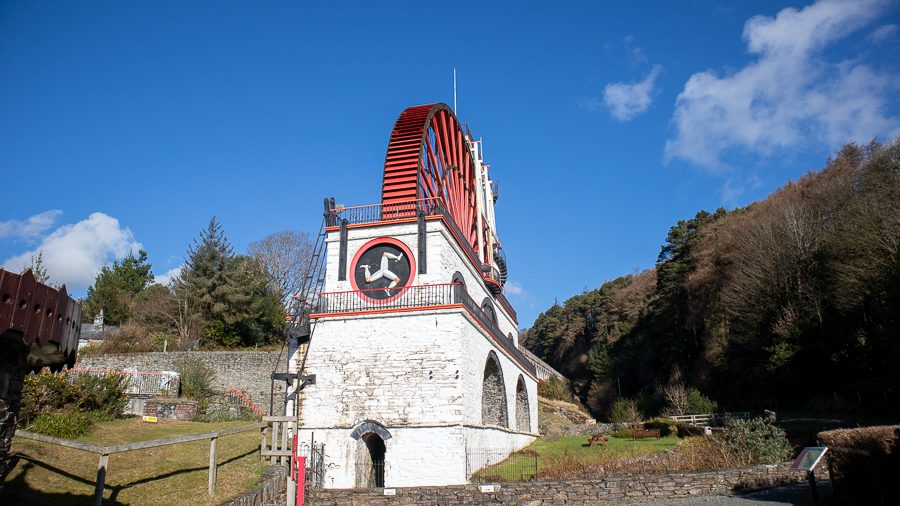 15+ Of The Best Things To Do On The Isle Of Man 7 Large red waterwheel with white brickwork and Isle of Man three-legged man on the front
