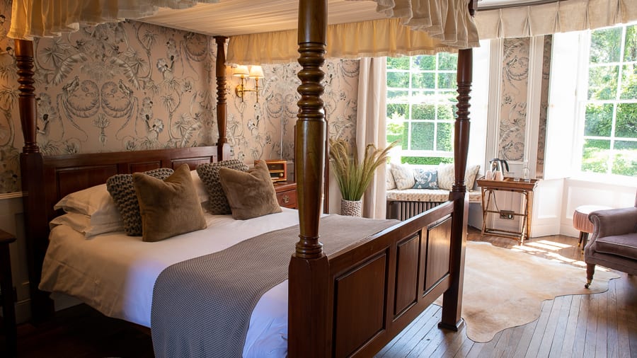 The Burleigh Court Hotel & Restaurant, Luxury in the Cotswolds 9 dark wood four-poster bed with mole coloured throw, caramel velvet cushions and pastel pink wallpaper with crested crane bird print in black, white and grey