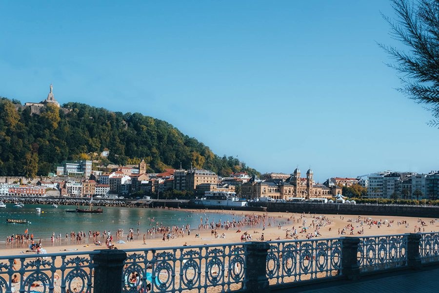 25 Amazing Destinations for Winter Sun around the World 24 ornate railings above a sandy beach with people on the sand and in the sea and the city of San Sebastian behind