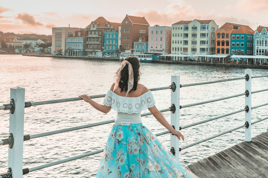 25 Amazing Destinations for Winter Sun around the World 8 pastel townhouses on the edge of the waters with a woman with her back to the camera wearing a white off-the-shoulder lacy top and turquoise floating floral skirt and a peach scarf in her hair