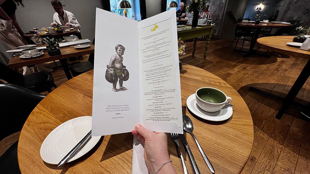 Luxury Stay at the Stunning Queensberry Hotel Bath 53 sitting at a round table looking at the menu with the list on the right and the drawn image of a young boy with boxing gloves on the left