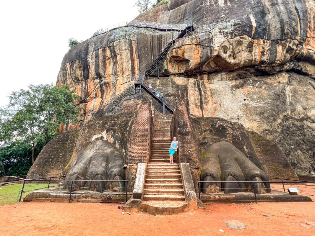 Climbing The Amazing Lion Rock In Sigiriya: The Ultimate Guide (2025) 11 Larch weating a blue t-shirt and shorts standing between the two great lion paws with the Lion Rock behind
