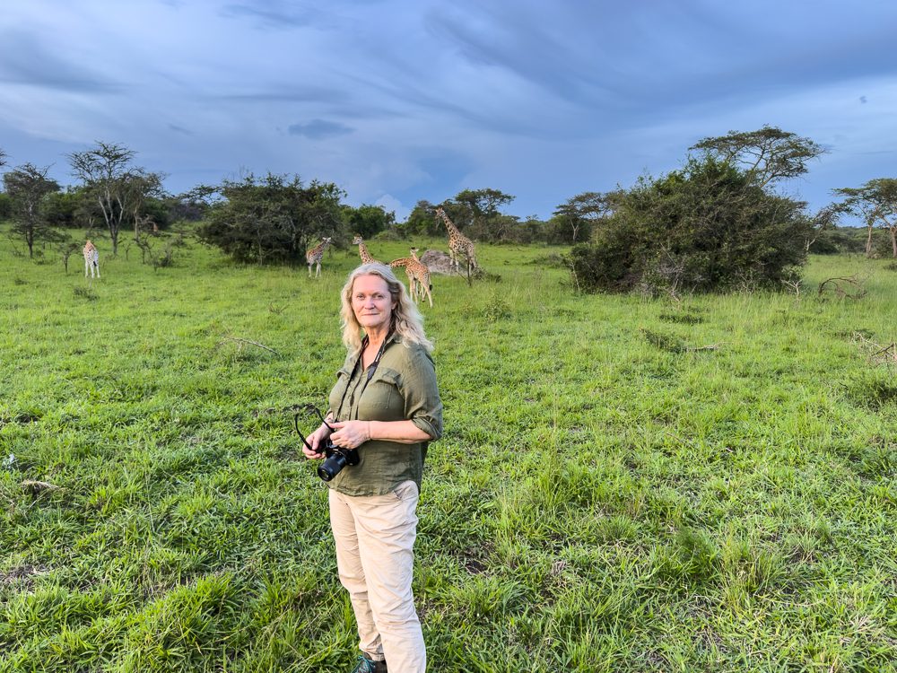 Larch wearing a green shirt and beige trousers and holding a camera with giraffes in the background
