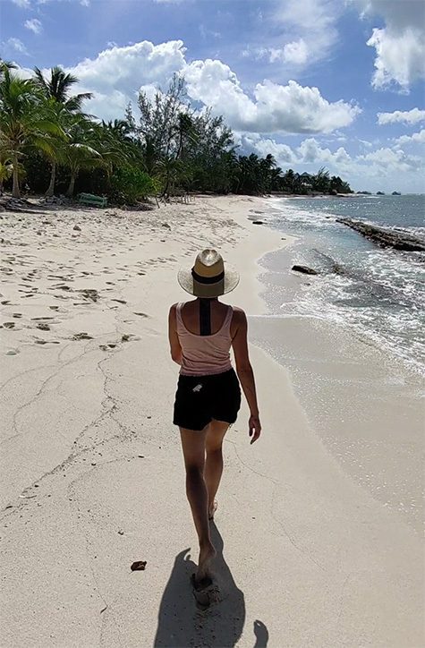 15 Amazing Hidden Gems To Discover In Grand Cayman 12 Woman walking on South Sound Beach in Grand Cayman wearing black shorts, pink sleeveless tshirt and straw hat, walking away from the camera.