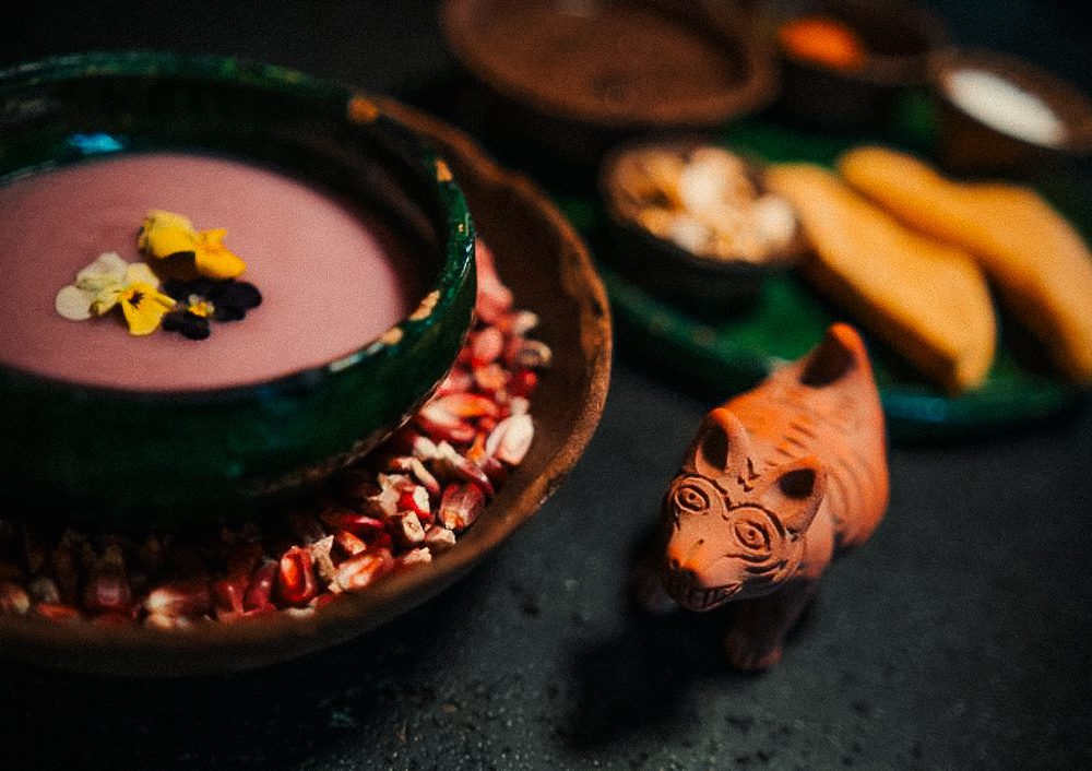 The Best Restaurants In Mexico City That Aren’t In The Usual Food Guides 6 A vibrant, artistic display of food featuring a colorful soup garnished with flowers, surrounded by corn kernels and a small decorative animal figure.